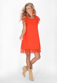 Rotes Kleid aus strukturiertem Stoff, mit einer Spitzenbordüre entlang des Ausschnitts und des Saums. Kombiniert mit beigen, im Western-Stil gehaltenen Ankle Boots.