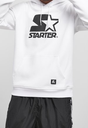 Osoba ubrana w białą bluzę z kapturem z czarnym logo „Starter” oraz czarne spodnie z białym napisem „Starter” po bokach, stojąca przodem.