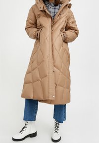 Tan pufferjas met een doorgestikt ontwerp, twee zijzakken en een hoge kraag. Gemaakt met blauwe jeans en witte veterschoenen.