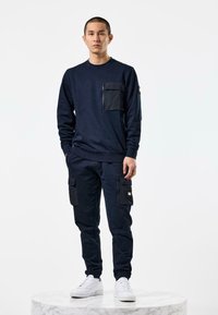 Námorníkový svetr s přední kapsou a odpovídající cargo kalhoty, obojí z měkké látky. Bílé tenisky dotvářejí outfit.