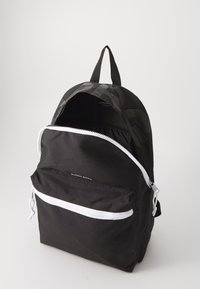 Mochila preta de tecido com um bolso frontal com fecho, fechos brancos e uma alça superior. Apresenta um padrão tonal sutil na parte de trás.