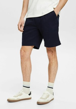 Laivastonsiniset puuvillashortsit sivutaskuilla ja suoralla helmalla, yhdistetty valkoisiin lenkkareihin ja raidallisiin sukkiin. Malli seisoo sivuttain neutraalia taustaa vasten.