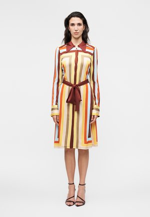 Mujer vestida con un vestido hasta la rodilla con rayas verticales y horizontales en marrón, naranja, amarillo y azul claro, atado con un cinturón de tela marrón.