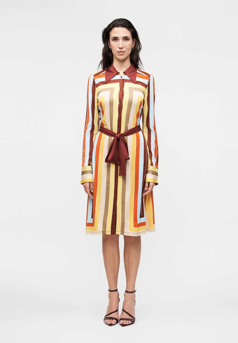 Γυναίκα που φοράει φόρεμα μήκους μέχρι το γόνατο με κάθετες και οριζόντιες ρίγες σε καφέ, πορτοκαλί, κίτρινο και ανοιχτό μπλε, δεμένο με καφέ υφασμάτινη ζώνη.