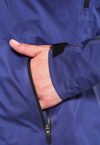 Main partiellement dans la poche zippée d'une veste bleue avec une sangle Velcro noire sur le poignet de la manche.