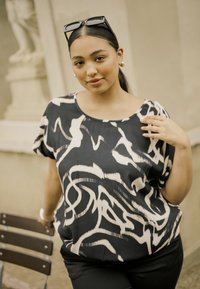 Jeune femme portant des lunettes de soleil sur la tête, vêtue d'un haut à motifs noir et blanc, se tenant à l'extérieur près d'une chaise et d'un mur en pierre.