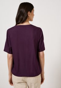 Blusa corta a maniche corte di colore viola con vestibilità ampia, caratterizzata da un collo rotondo e dettagli con bottoni sulle maniche. Tessuto liscio con un leggero lucido.