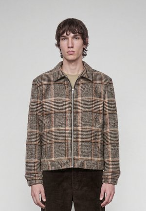 Ung mand med kort krøllet hår iført en brun og beige ternet lynlåstrøje og mørkebrune bukser mod en ensfarvet baggrund.