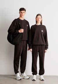 Čierna súprava mikiny a teplákov z mäkkého materiálu. Oba kúsky majú malú, diamantovo tvarovanú nášivku. Oblečenie je doplnené bielymi teniskami.