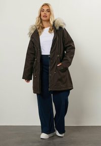 Kvinna med långt blont hår som bär en mörkbrun parkas med pälsmössa, vit t-shirt, vida mörka jeans och vita sneakers, som står på en grå golv.