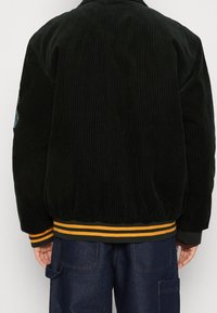 Czarna kurtka bomber wykonana z sztruksu z żółto-zielonymi paskami na ściągaczu, z naszywką na lewym rękawie i luźnym krojem.