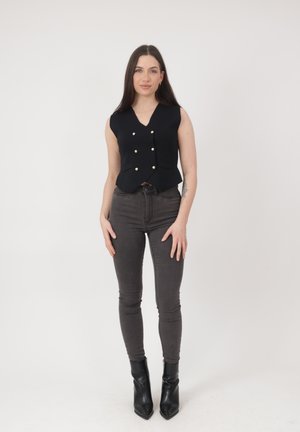 Kobieta stojąca, ubrana w bezrękawnik w czarnym kolorze z guziczkami, ciemne dopasowane dżinsy oraz czarne botki na kostkę, na tle jasnego, jednolitego tła.
