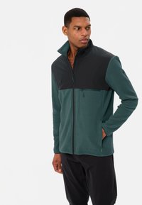 Giacca in fleece con zip completa, caratterizzata da un corpo verde scuro e pannelli sulle spalle neri, tessuto testurizzato e tasca laterale.