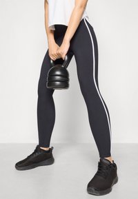 Black leggings with white side stripes, paired with black athletic shoes. The individual is holding a black kettlebell.