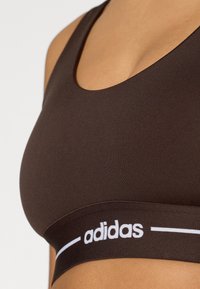 Hnedá športová podprsenka z hladkej, elastickej tkaniny; má výstrih do „véčka“ a biely pás s logom pozdĺž spodnej časti.