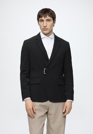 Jeune homme aux cheveux courts et bruns porte un blazer noir avec une boucle argentée, une chemise blanche boutonnée et un pantalon beige, faisant face à l'avant.