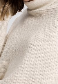 Gros plan sur une personne portant un pull en coton écru à col roulé avec des points texturés, les cheveux jusqu'à l'épaule visibles.