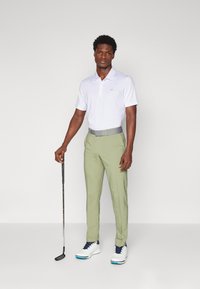 Bela polo majica s ovratnikom, svetlo zelene hlače in bele golf čevlje z modrimi akcenti. Model drži golf palico in stoji pred nevtralno ozadje.