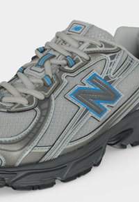 Γκρι και μπλε αθλητικό παπούτσι New Balance με υφασμάτινη υφή, χοντρή σόλα και λεπτομερή κορδόνια σε κοντινή λήψη.