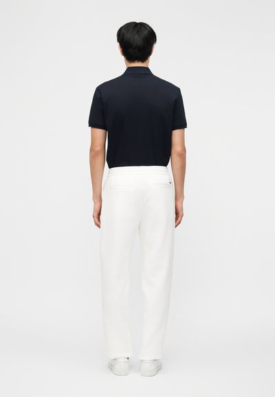 Άνδρας που στέκεται με την πλάτη γυρισμένη, φοράει μαύρο κοντομάνικο πόλο, λευκό παντελόνι με φαρδιά πόδια και λευκά αθλητικά παπούτσια σε απλό φόντο.