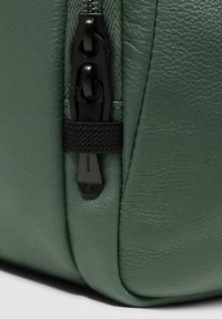 Angolo di borsa in pelle verde con superficie testurizzata, dotata di cerniera nera e linguetta. Il design include un accento in tessuto intrecciato vicino alla cerniera.