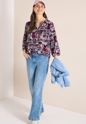 Donna sorridente che indossa un berretto rosa, una camicetta a fantasia e jeans azzurri, con una giacca di denim piegata nella mano sinistra.