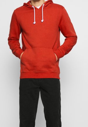 Uomo che indossa una felpa rossa con cappuccio e lacci bianchi, pantaloni di velluto a coste neri, mani nelle tasche della felpa.