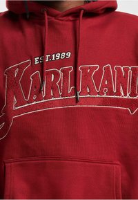 Rød hettegenser med "KARL KANI" i store, teksturerte hvite bokstaver med "EST.1989" over. Inkluderer frontlomme og justerbare snøring.