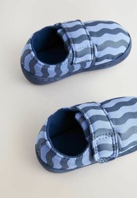 Blå slip-on skor med ett vågigt randigt mönster i ljus och mörk blå, med en kardborreband och ett mjukt textilövre.