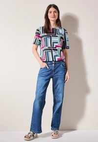 Haut multicolore à manches courtes avec des motifs géométriques, associé à un jean en denim bleu à jambes larges et des sandales argentées.