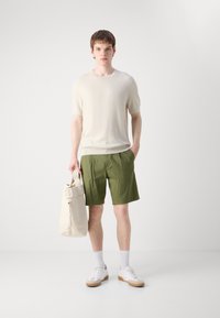 Maglioncino lavorato a maglia crema a maniche corte, pantaloni shorts verde oliva, borsa beige a spalla, sneakers bianche e calze bianche, posizionati su uno sfondo neutro.