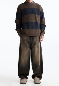 Pull en tricot rayé marron et bleu, avec poignets et ourlet côtelés. Porté avec un jean marron délavé et ample, ainsi que des chaussures grises à enfiler.