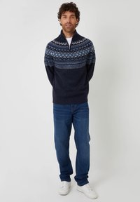 Granatowy sweter z wysokim kołnierzem i wzorzystym karczkiem w odcieniach niebieskiego. Noszony z niebieskimi dżinsami i białymi trampkami. Model stoi z dłońmi splecionymi.