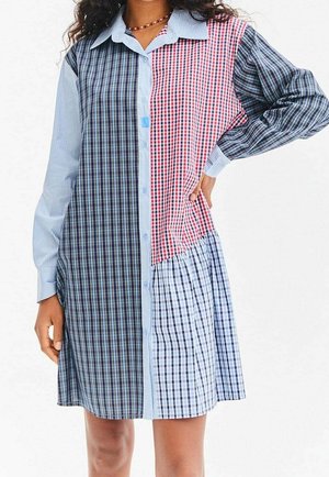 Femme portant une robe-chemise à manches longues, à carreaux multi-patterns avec des sections bleues, rouges et noires, une main sur la hanche, sur fond blanc.