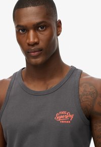 Hombre con una camiseta sin mangas de color gris oscuro con el logo naranja "Tokyo Superdry" y texto en japonés, mostrando tatuajes en el hombro derecho y la parte superior del brazo.