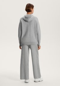 Siva pletena kapucha s široko silhueto, z žametenim robom in stranskimi šivi. V kombinaciji s sivimi širokimi hlačami, ki imajo gladko teksturo.