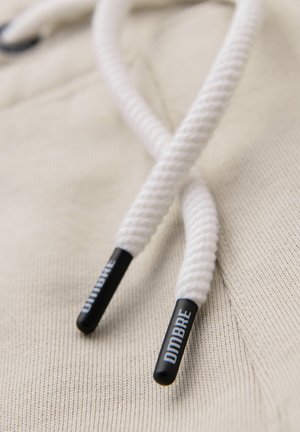 Nahaufnahme von weißen Kapuzenkordeln mit schwarzen Plastiktips, die mit "DIMBRE" beschriftet sind, auf beigem strukturiertem Stoff.