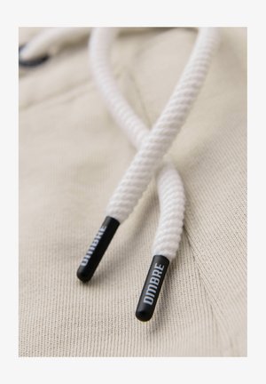 Gros plan sur les cordons de capuche blancs avec embouts noirs en plastique portant l'inscription "DIMBRE" sur un tissu beige texturé.