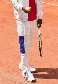 Hvit idrettsjakke med en blå panel, rød skjorte, holder en tennisball i den ene hånden og en racket i den andre. Hvite tennissko.
