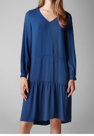 Woman wearing a long-sleeve, knee-length, loose-fitting blue dress with a V-neck and gathered skirt detail.