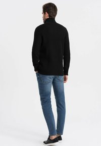 Maglione nero a maglia con colletto alto, abbinato a jeans slim-fit blu e scarpe slip-on nere. Texture visibili e vestibilità rifinita.