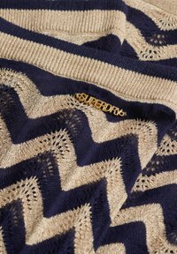 Gebreide trui met een chevronpatroon in marineblauw en goud, met een gestructureerd oppervlak en een zichtbare 'Superdry' logo-accessoire.