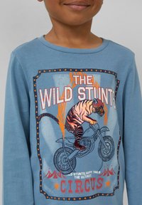 Kind met een blauwe shirt met een afbeelding van een tijger in een helm die een motorstunt uitvoert door een vlammenring, circus thema.