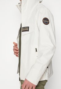White lightweight jacket with a hood, zip closure, and a patch on the sleeve. Features side pockets and adjustable cuffs.