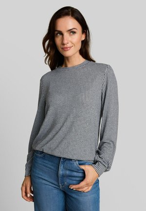 Vrouw met schouderlang bruin haar, draagt een zwart-wit patroon lange mouwen top en blauwe spijkerbroek met één hand in de zak.