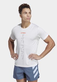 Camiseta atlética blanca con paneles laterales transpirables, que presenta un diseño gráfico vertical en gris y un logo en naranja. Combinada con pantalones cortos azules.