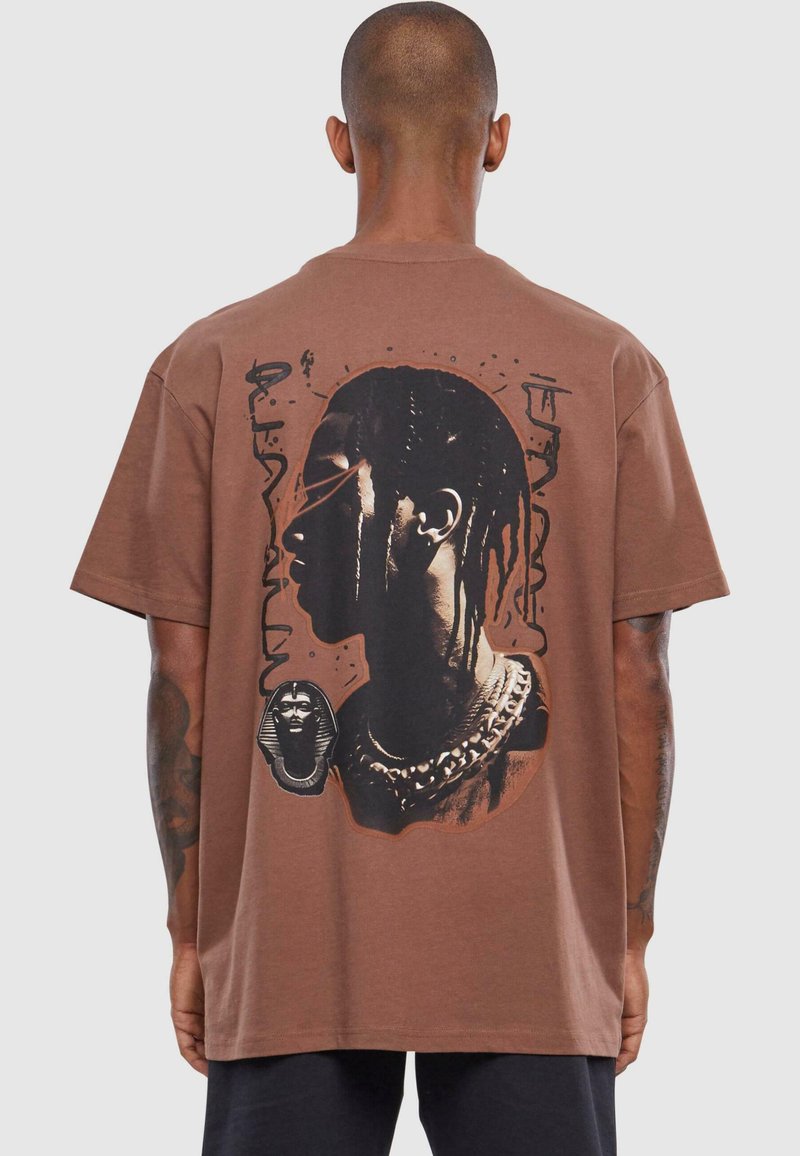 Brun bomulds T-shirt med et stort grafisk motiv på ryggen, der viser en profileillustration, kæder og egyptiske motiver. Korte ærmer.