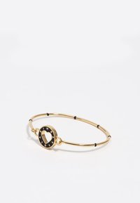 Pulsera de metal dorado con un charm circular que presenta un corte en forma de corazón, acentos de esmalte negro y cuentas negras alternas a lo largo de la banda.