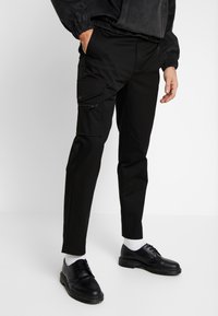 Černé cargo kalhoty se slim fit, bočními kapsami na zip a hladkým povrchem, spárované s černými botami a bílými ponožkami.