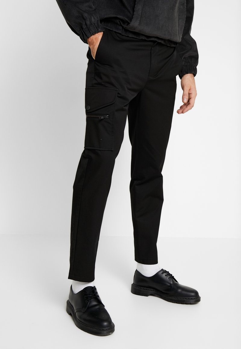 Černé cargo kalhoty se slim fit, bočními kapsami na zip a hladkým povrchem, spárované s černými botami a bílými ponožkami.